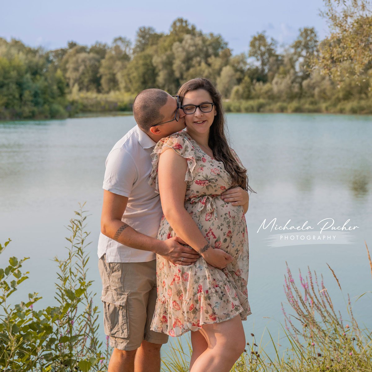 Schwangere Frau in einem geblümten Kleid steht am See, während ihr Partner sie liebevoll von hinten umarmt und auf die Wange küsst.